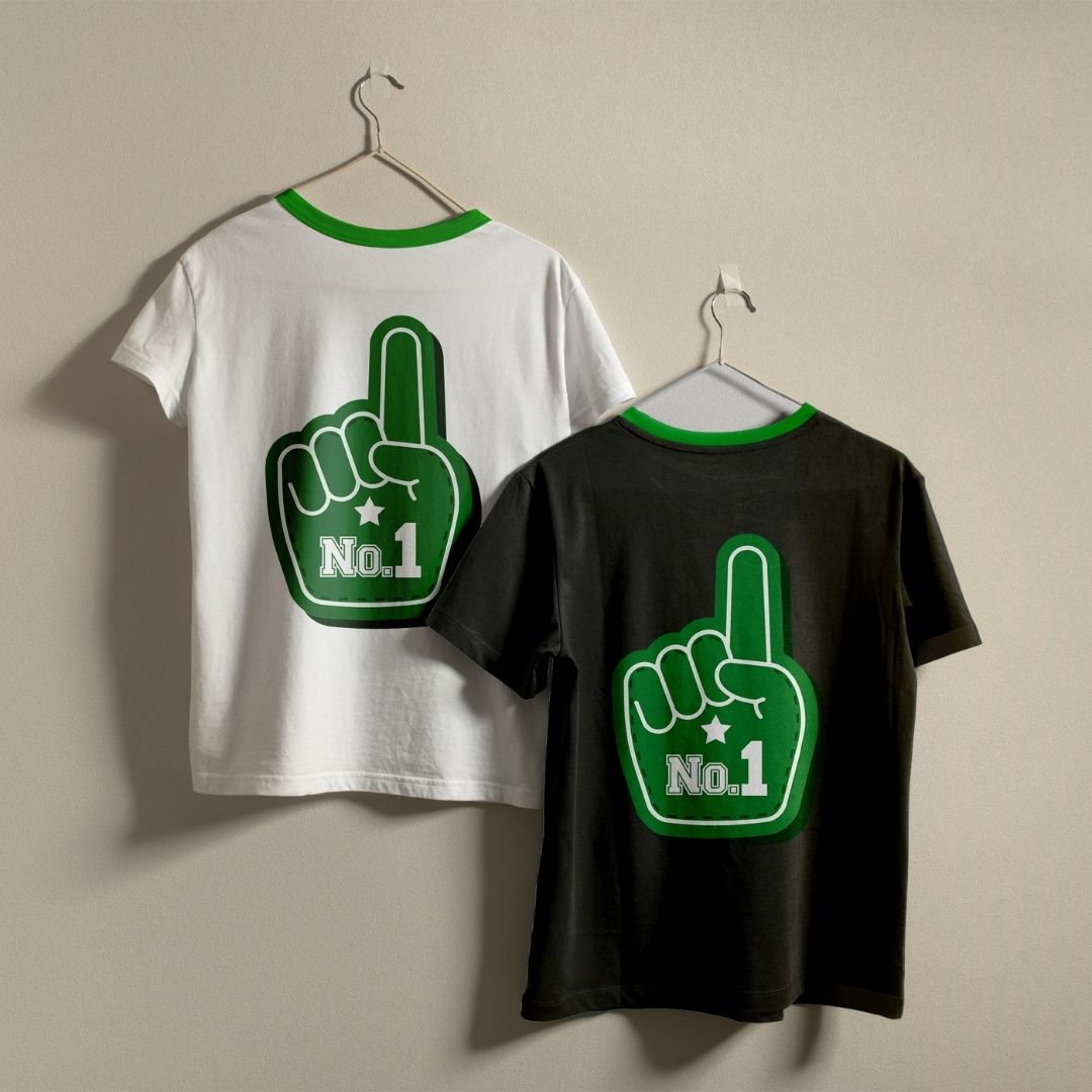 Two ringer t-shirts with green neck trims, one white and one black, hanging on metal hangers against a beige wall. Both shirts feature a large green foam finger graphic on the back with 'No.