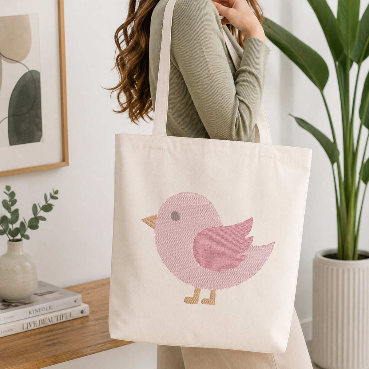 A person wearing a sage green ribbed top carries a cream canvas tote bag with a pink bird graphic. The scene is set in a bright, modern room with a wooden table, books, a small plant, and a large green potted plant in the background.