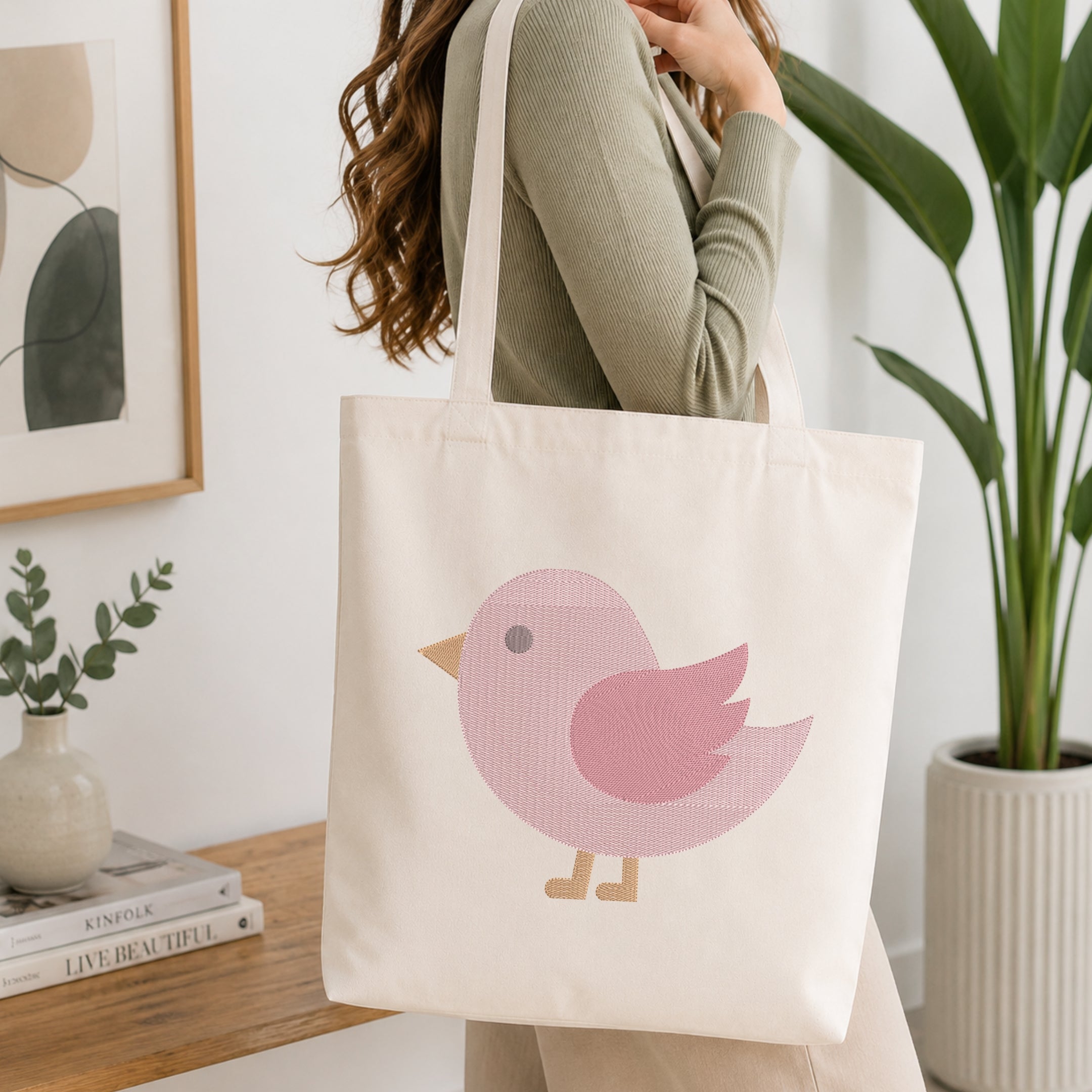 A person wearing a sage green ribbed top carries a cream canvas tote bag with a pink bird graphic. The scene is set in a bright, modern room with a wooden table, books, a small plant, and a large green potted plant in the background.