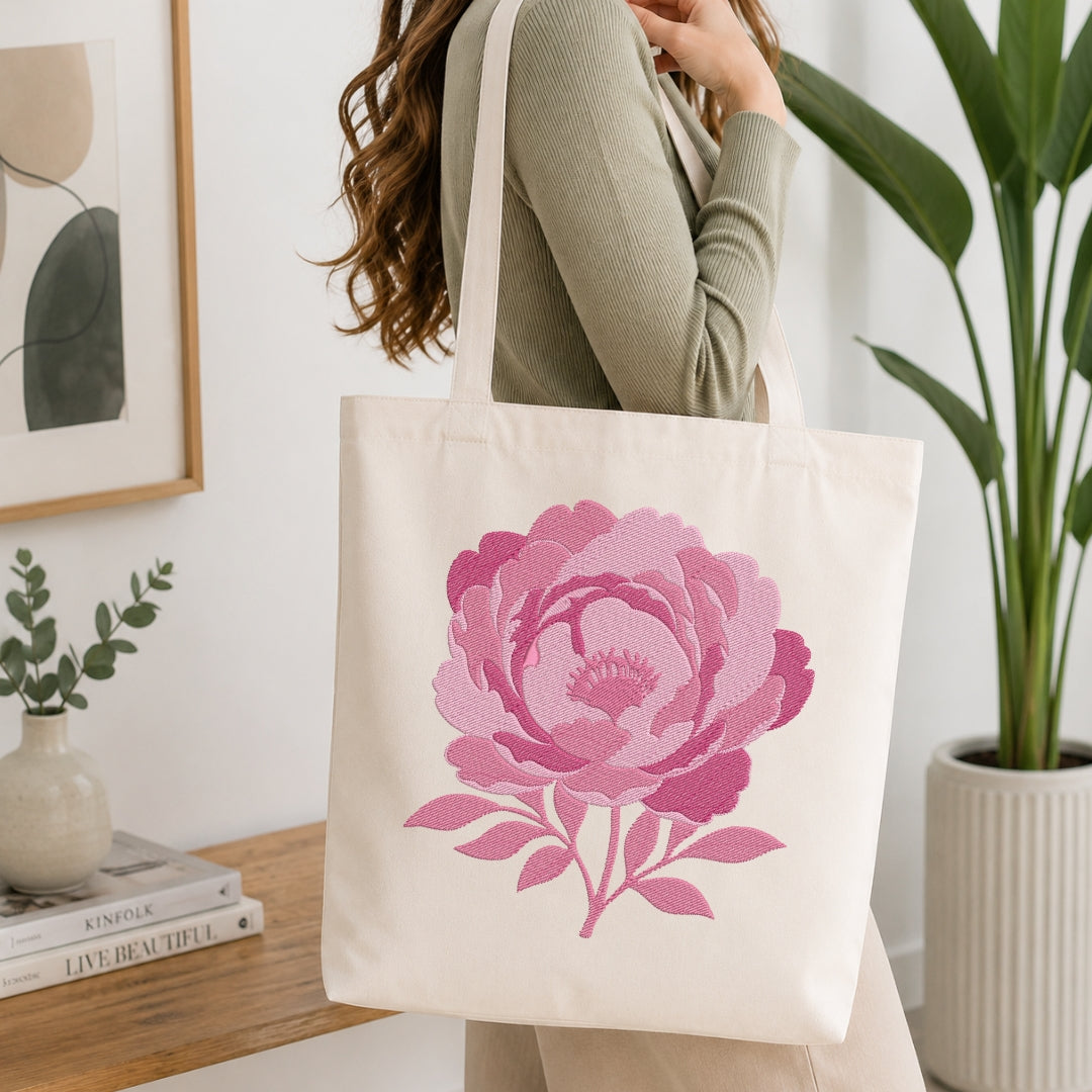 A woman wearing a sage green ribbed sweater carries a cream canvas tote bag with a large pink peony flower print. The scene is set in a bright, modern room with a wooden table, books, a small vase, and a potted green plant.