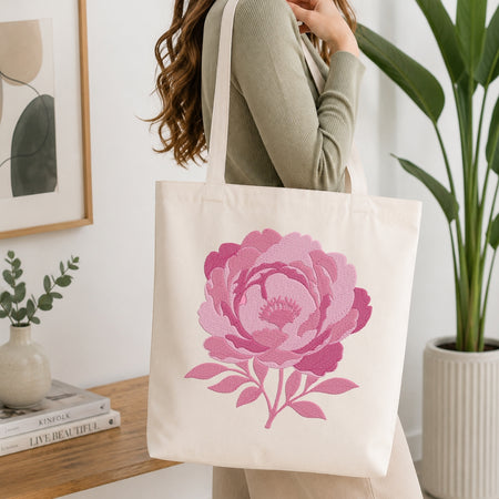 A woman wearing a sage green ribbed sweater carries a cream canvas tote bag with a large pink peony flower print. The scene is set in a bright, modern room with a wooden table, books, a small vase, and a potted green plant.