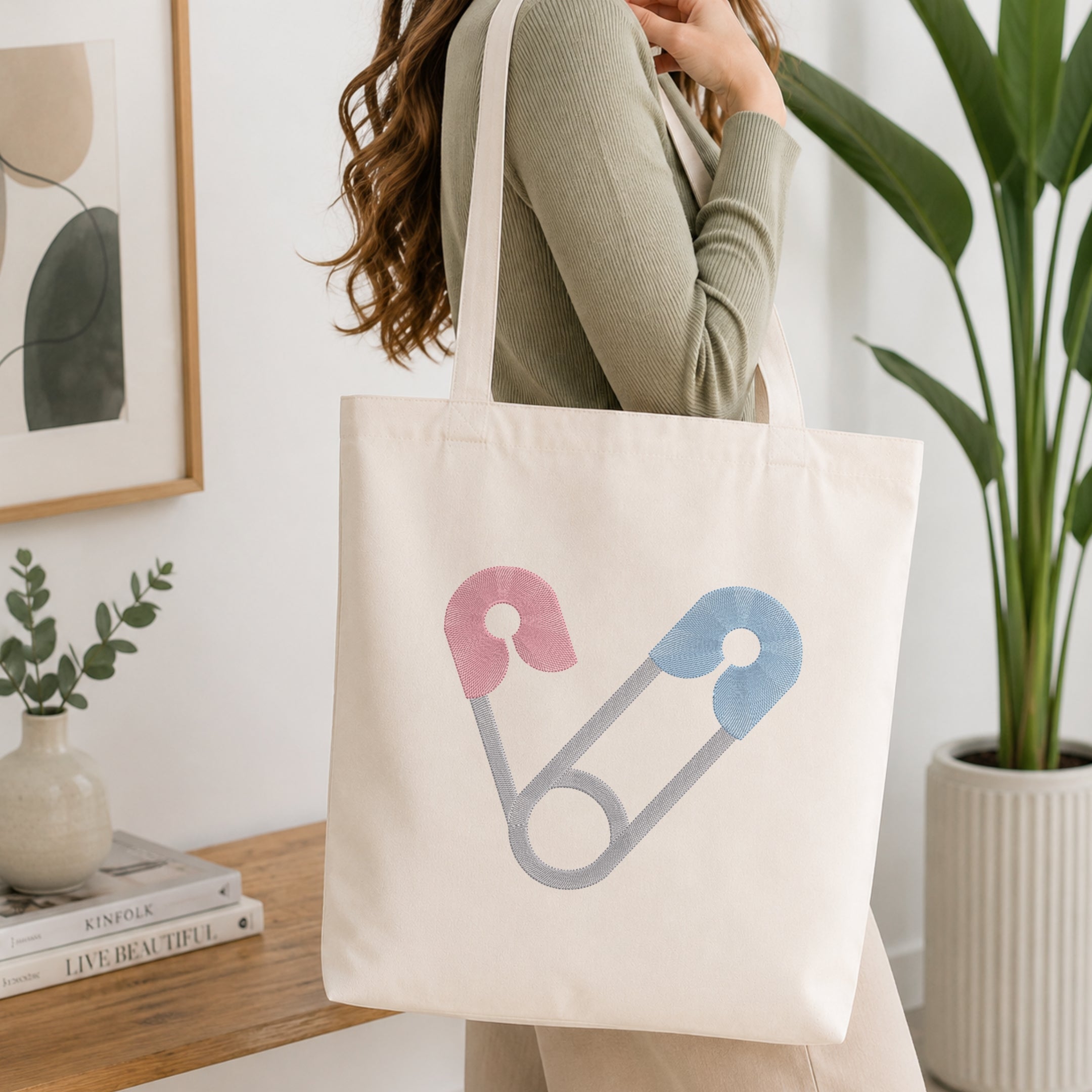 A woman wearing a sage green ribbed long-sleeve top carries a cream canvas tote bag with a safety pin graphic in pink, blue, and gray. The background is a bright, modern room with a wooden shelf, framed art, and a potted plant.