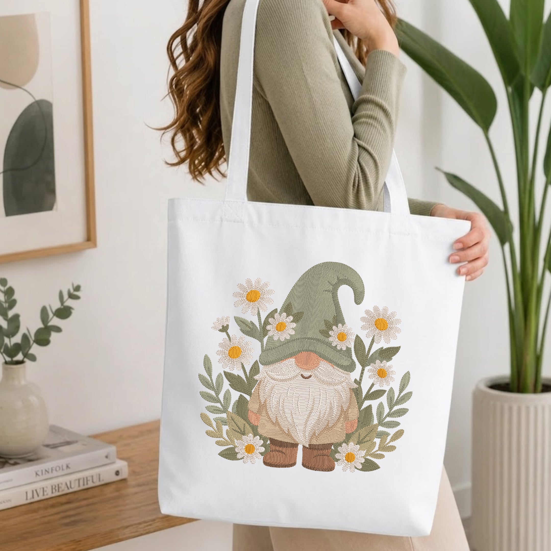A woman carries a white canvas tote bag featuring a cute gnome illustration surrounded by daisies and green leaves. The scene is set in a bright, modern room with a wooden table, books, and a potted plant.