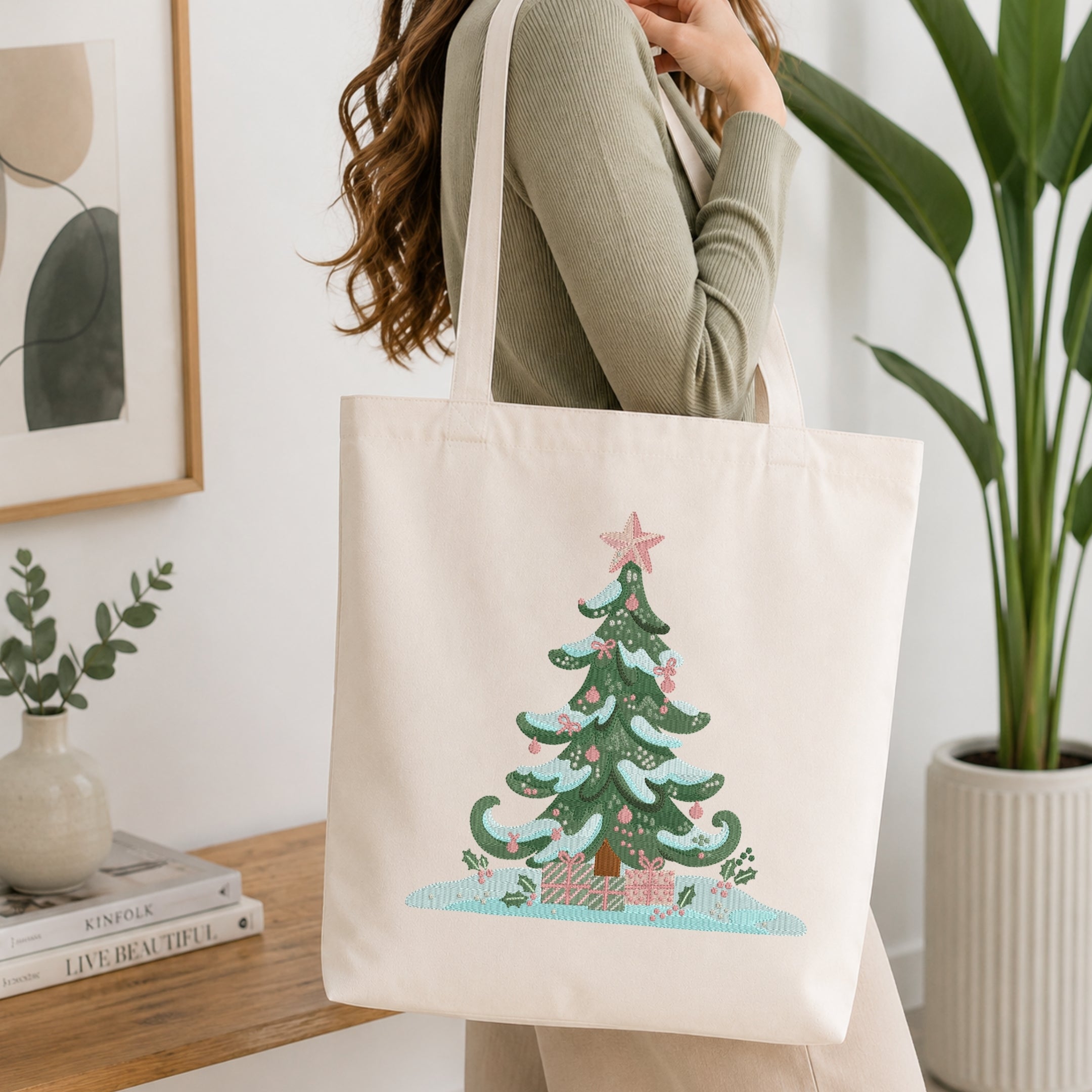A woman wearing a sage green ribbed sweater carries a cream canvas tote bag with a printed Christmas tree design. The tote bag features a green tree with blue snow accents, pink ornaments and star, and a pink gift box at the bottom.