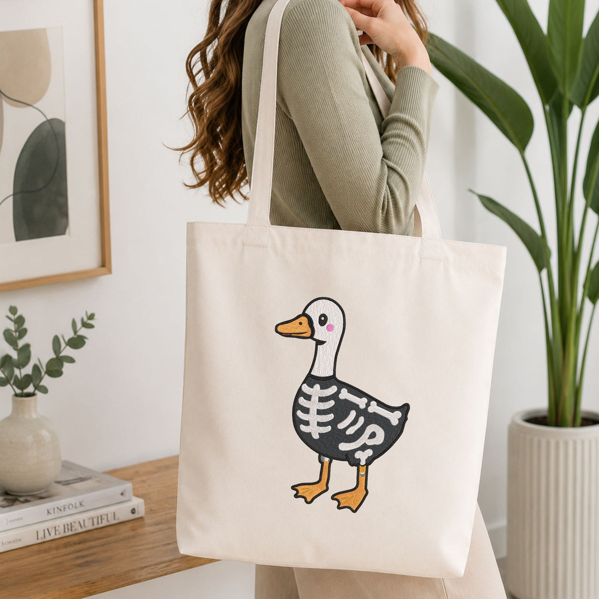 Woman carrying a white natural canvas tote bag featuring a cute skeleton duck Halloween machine embroidery design in a bright, modern interior setting
