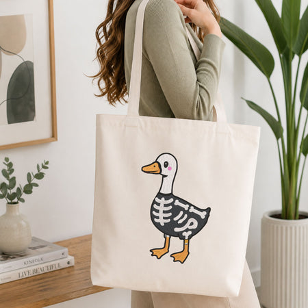 Woman carrying a white natural canvas tote bag featuring a cute skeleton duck Halloween machine embroidery design in a bright, modern interior setting