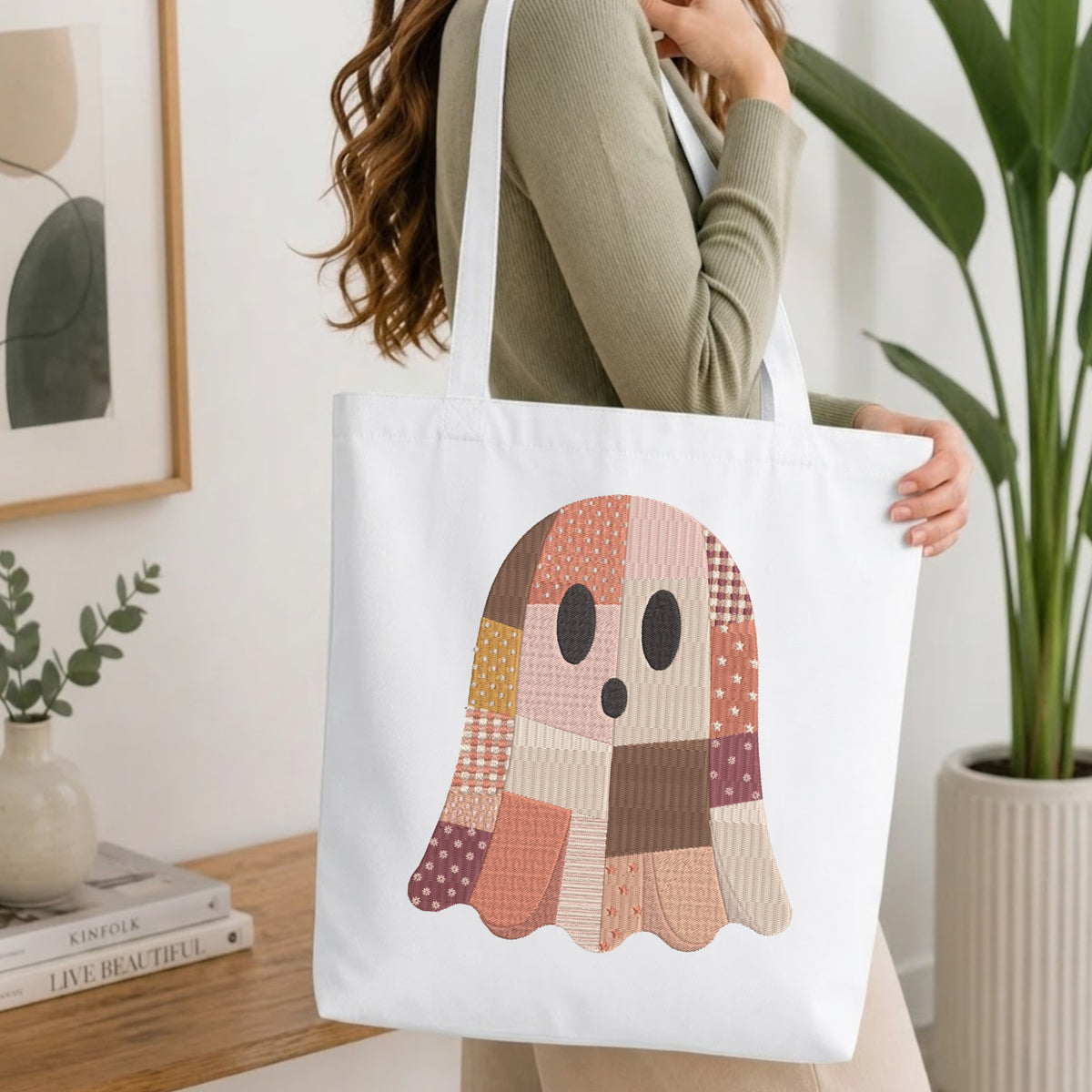 Woman carrying a natural canvas tote bag featuring an embroidered patchwork Halloween ghost design in a bright, modern interior setting.