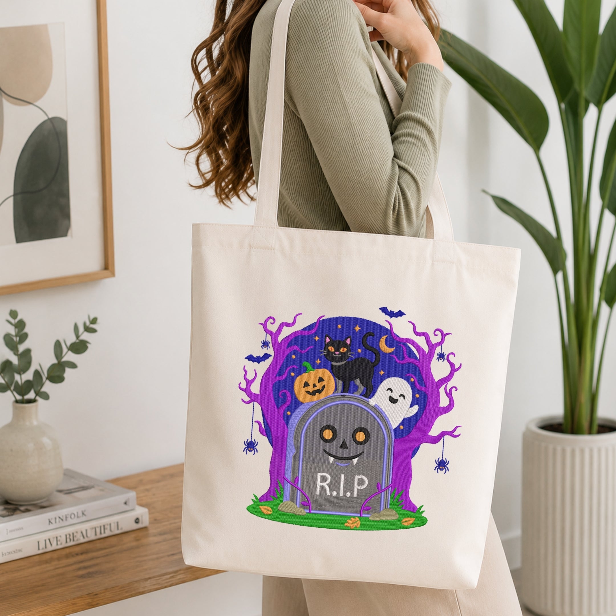 A woman wearing a sage green ribbed long-sleeve top carries a cream canvas tote bag with a colorful Halloween graphic. The graphic features a smiling tombstone with 'R.