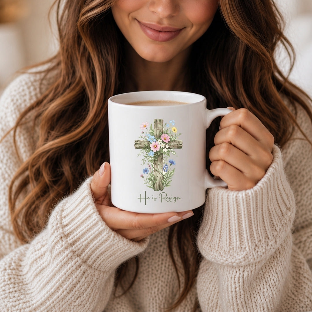 A woman with long brown hair holding a white ceramic mug featuring a floral cross design and the text "He is Resign".