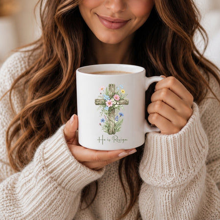A woman with long brown hair holding a white ceramic mug featuring a floral cross design and the text "He is Resign".