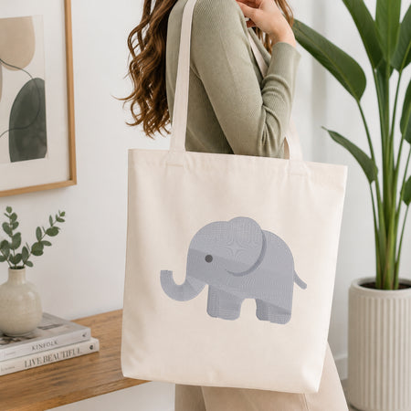 A woman wearing a sage green ribbed top carries a cream canvas tote bag with a light gray elephant graphic printed on the front, set in a bright, modern room with a wooden table, framed art, and a potted plant.