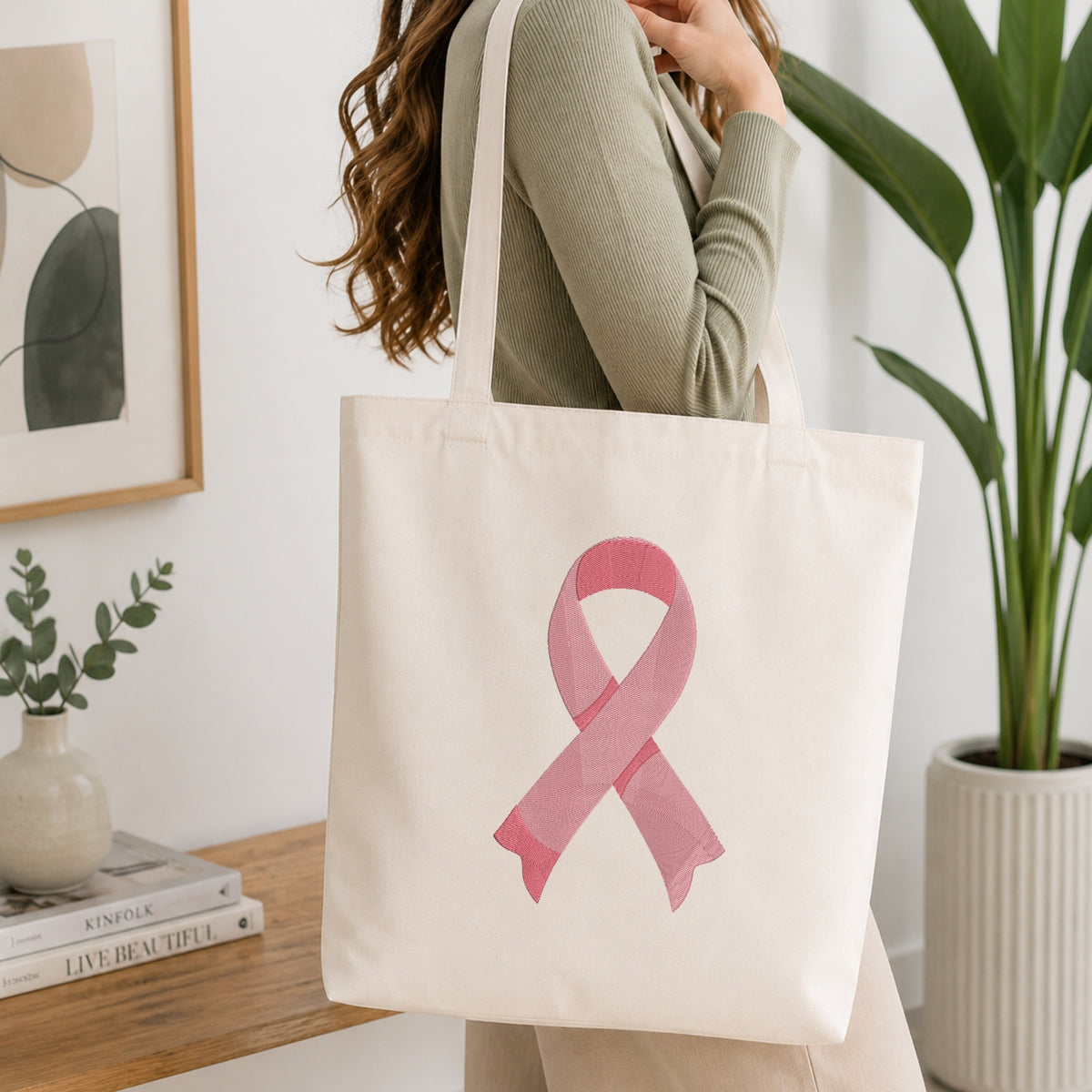 A woman wearing a sage green ribbed long-sleeve top carries a cream canvas tote bag with a pink watercolor ribbon design on the front, set in a bright, modern room with a wooden table, framed art, and a potted plant.