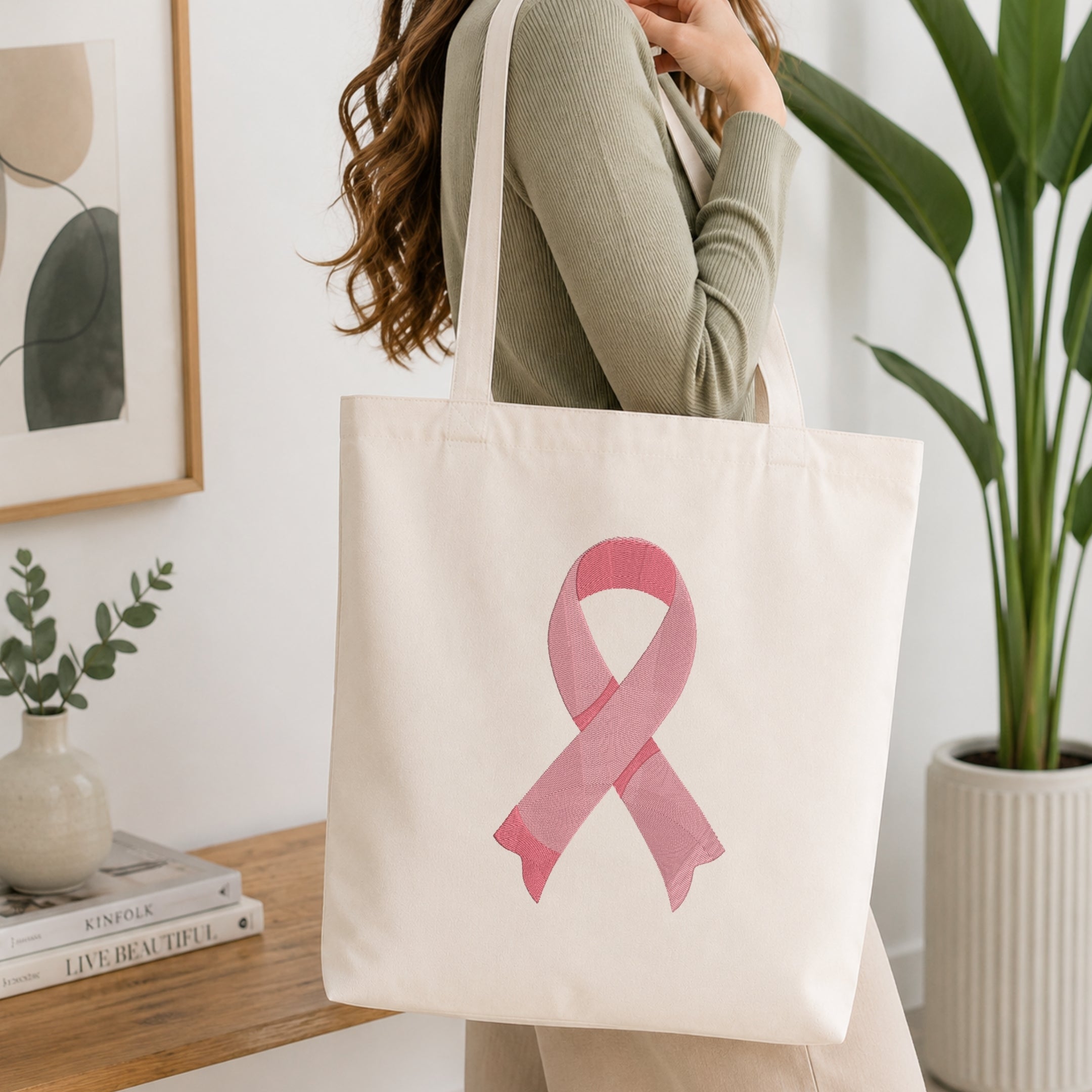 A woman wearing a sage green ribbed long-sleeve top carries a cream canvas tote bag with a pink watercolor ribbon design on the front, set in a bright, modern room with a wooden table, framed art, and a potted plant.