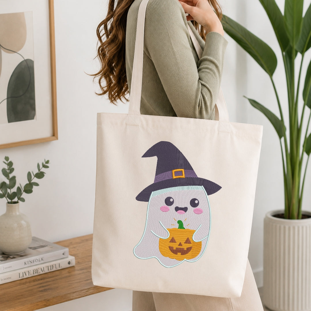 An off-white canvas tote bag with a cute embroidered ghost and pumpkin design, carried by a woman in a ribbed sage green top in a bright, modern room.