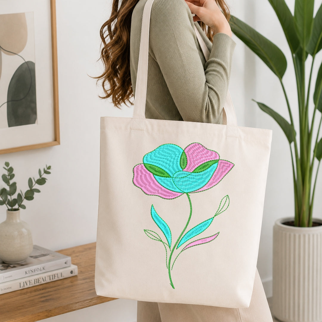 A woman wearing a sage green ribbed sweater carries a cream canvas tote bag with a colorful embroidered flower design in pink, teal, and green, set against a bright, modern interior background with a wooden table and potted plants.
