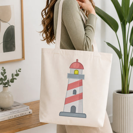 A woman wearing a sage green ribbed sweater carries a cream canvas tote bag with an embroidered lighthouse design. The lighthouse features red and white stripes, a red roof, a yellow window, and a gray base.