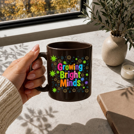 A hand holds a dark brown ceramic mug with the colorful text 'Growing Bright Minds' surrounded by star and swirl patterns. The mug is shown in a warm, sunlit room setting on a windowsill next to a potted plant and a candle.