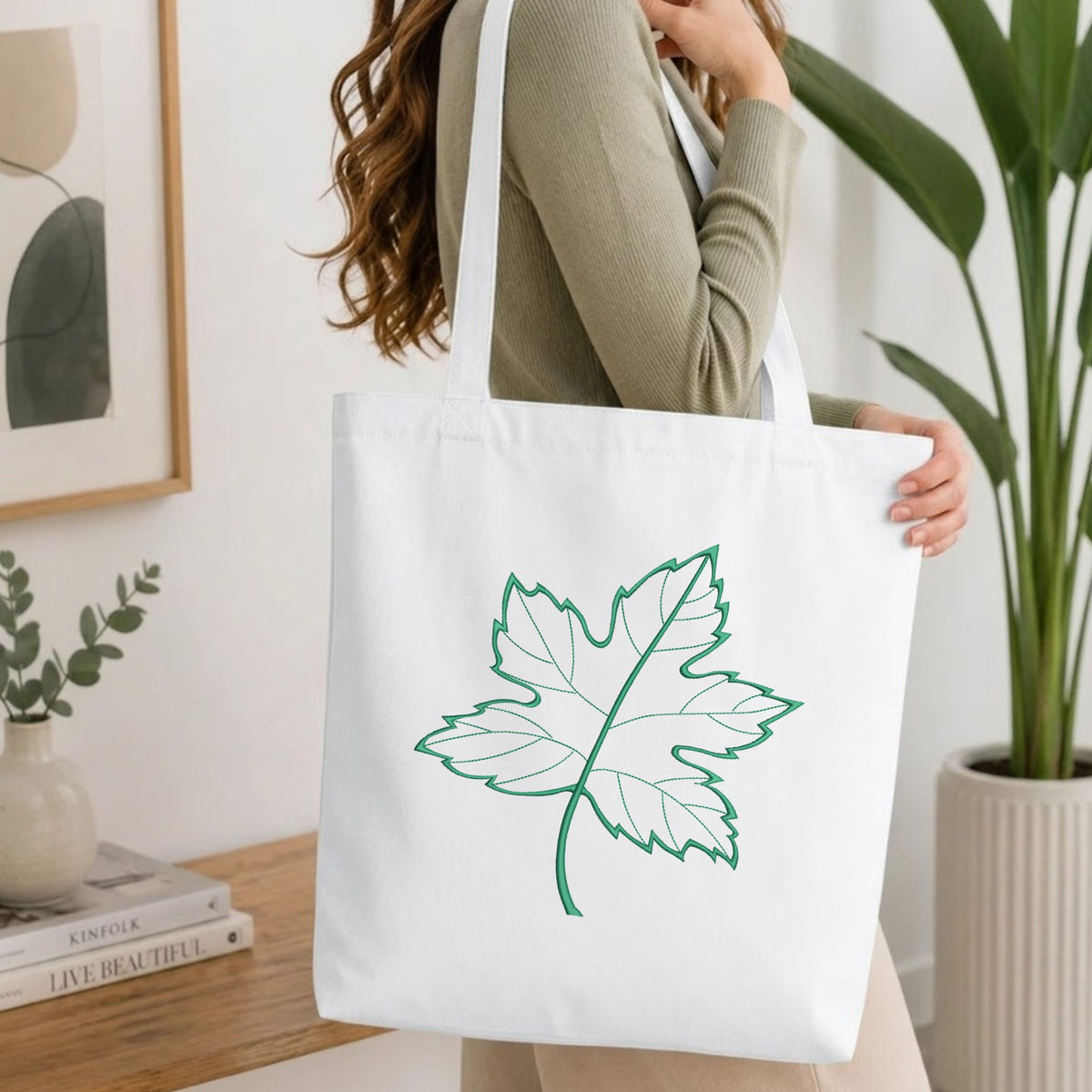 Woman carrying a white natural canvas tote bag featuring an embroidered green maple leaf design in a bright, modern interior setting