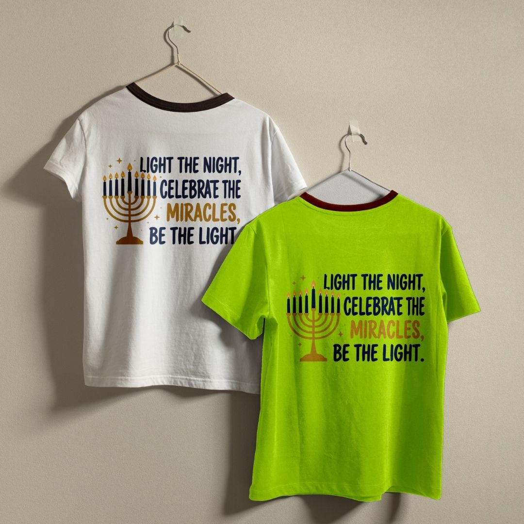 Two ringer t-shirts hanging on metal hangers against a beige wall. The left shirt is white with a dark red collar and features a Hanukkah-themed graphic with a gold menorah and the text 'LIGHT THE NIGHT, CELEBRATE THE MIRACLES, BE THE LIGHT.