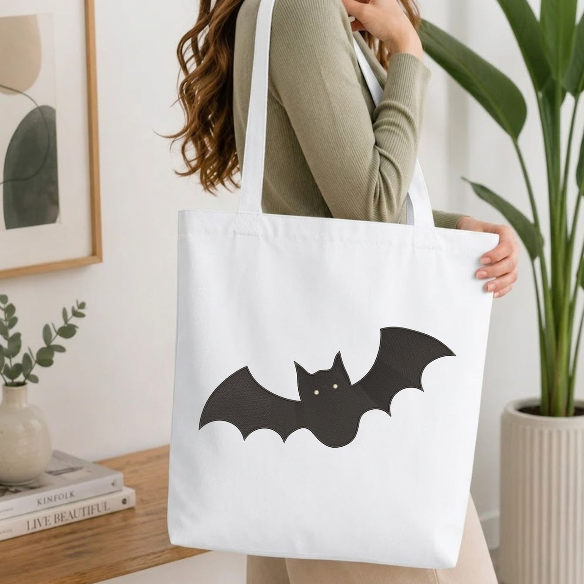 Woman carrying a white natural canvas tote bag featuring an embroidered spooky cute black bat design in a bright, modern interior setting.