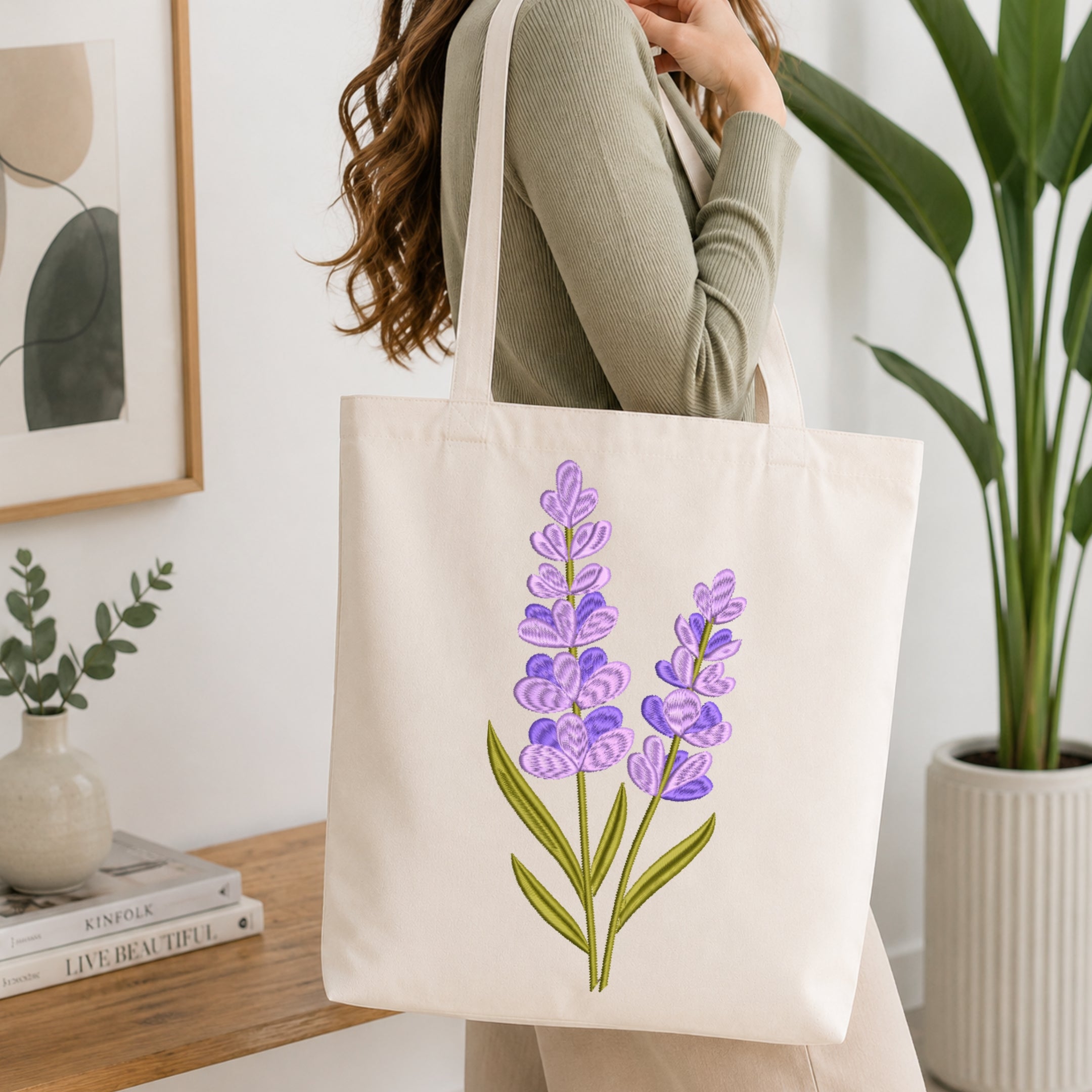 A woman wearing a sage green ribbed sweater carries a cream canvas tote bag with a purple and green lavender floral print. The scene is set in a bright, modern room with a wooden table, books, a small plant, and a large green potted plant in the background.