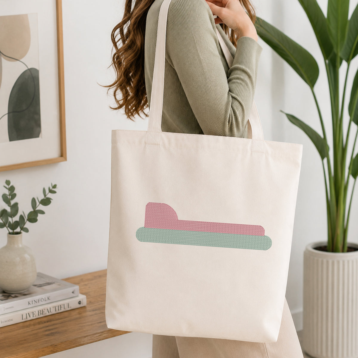A woman wearing a sage green ribbed top carries a cream canvas tote bag with a tan bone outline design, shown in a bright, modern room with a wooden table, framed art, and a potted plant.