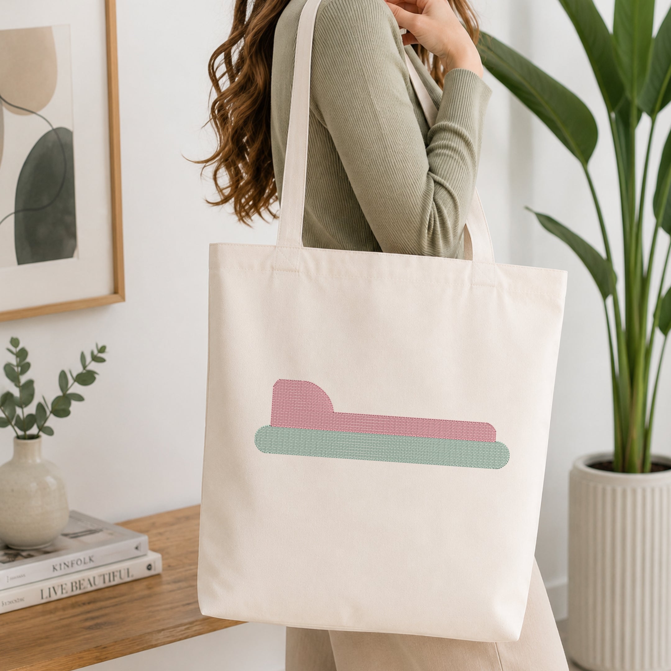 A woman wearing a sage green ribbed top carries a cream canvas tote bag with a tan bone outline design, shown in a bright, modern room with a wooden table, framed art, and a potted plant.