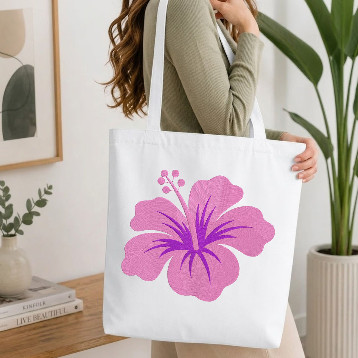 A woman wearing a sage green ribbed long-sleeve top carries a white canvas tote bag featuring a large pink hibiscus flower graphic. The setting is a bright, modern room with a wooden table, books, a framed print, and a potted green plant.