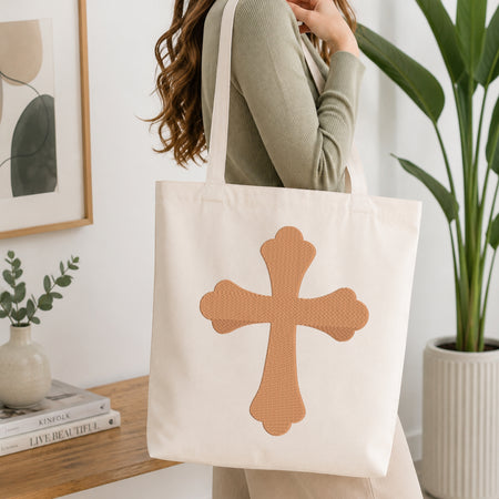 A woman wearing a sage green ribbed long-sleeve top carries a cream canvas tote bag with a large tan textured cross on the front, set in a bright, modern room with a wooden table, framed art, and a potted plant.