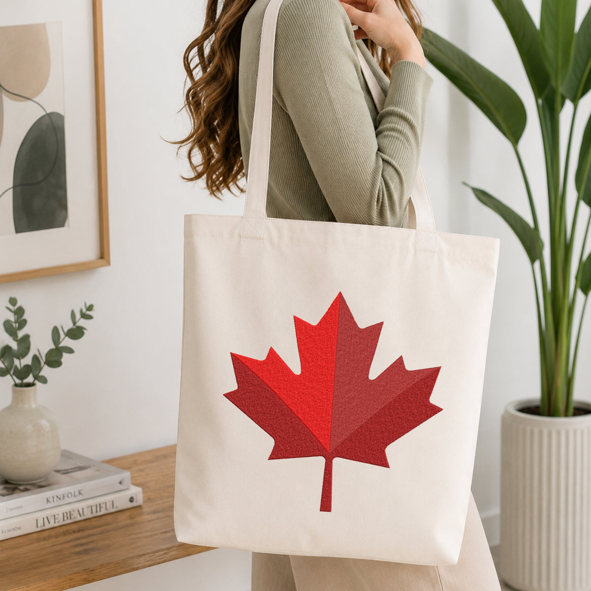 A woman wearing a sage green ribbed sweater carries a cream canvas tote bag with a large red maple leaf graphic on the front, set in a bright, modern room with a wooden table, books, and a potted plant.