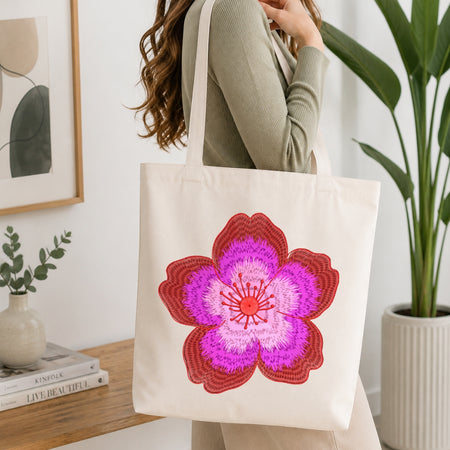 A woman wearing a sage green ribbed sweater carries a cream canvas tote bag with a large, vibrant pink and red floral patch on the front, set in a bright, modern room with a wooden table and potted plants.