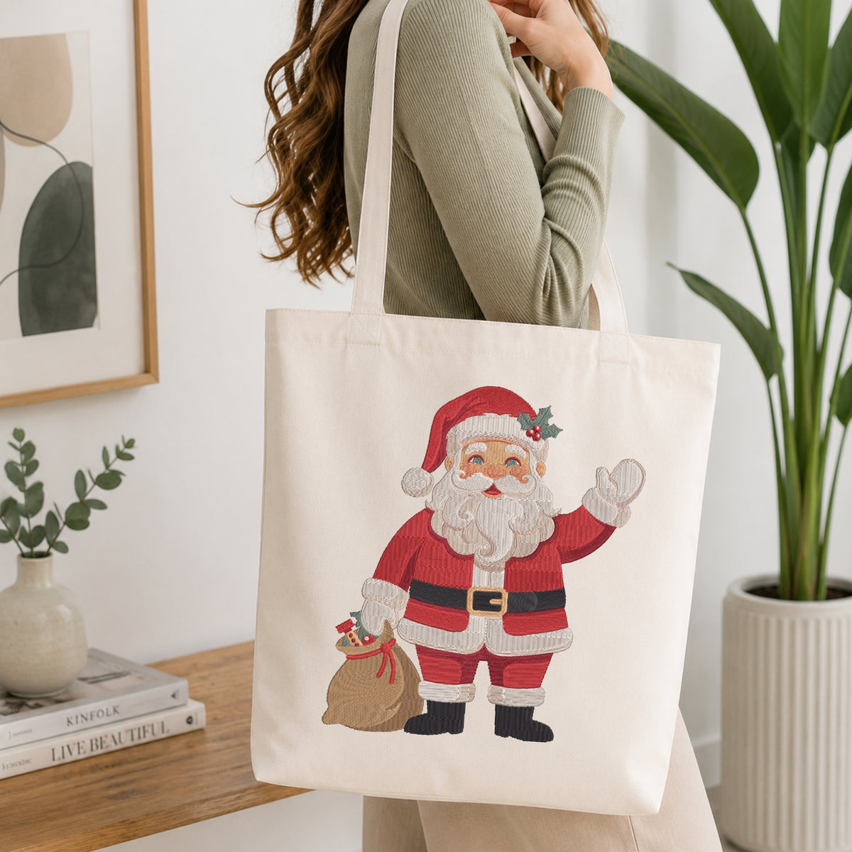 A woman wearing a sage green ribbed sweater carries a cream canvas tote bag featuring a colorful embroidered Santa Claus design. The scene is set in a bright, modern room with a wooden table, books, a framed print, and a potted plant.