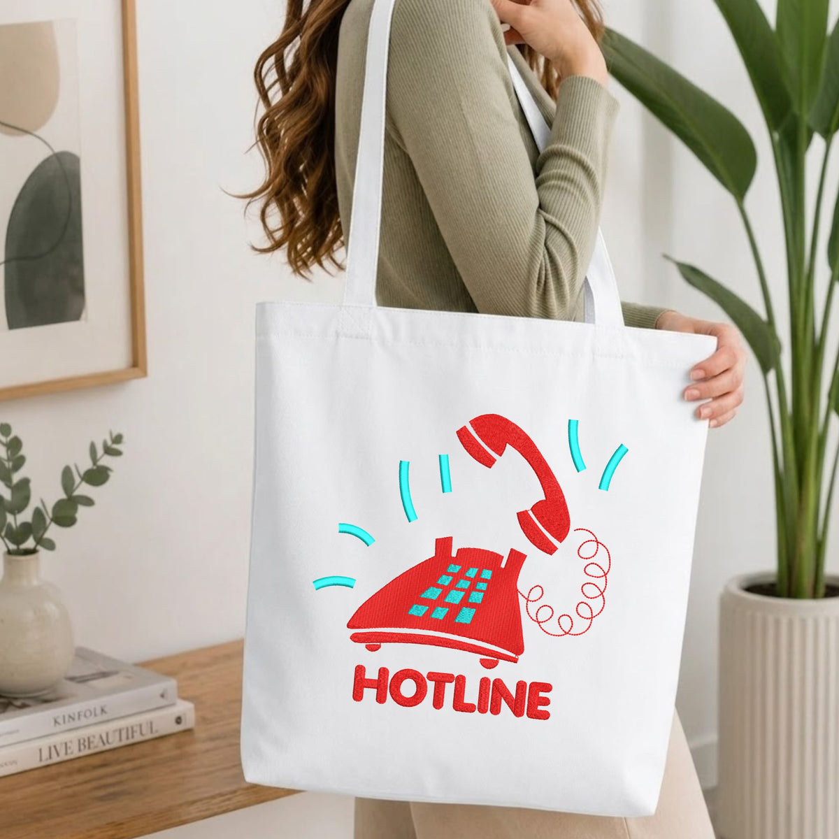 Woman carrying a white natural canvas tote bag featuring an embroidered red vintage telephone design in a bright, modern interior setting
