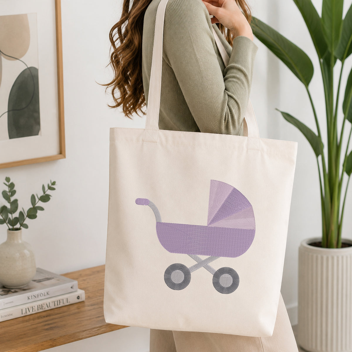 A woman wearing a sage green ribbed top carries a cream canvas tote bag featuring a purple and gray baby stroller graphic, set in a bright, modern room with a wooden table, framed art, and a potted plant.