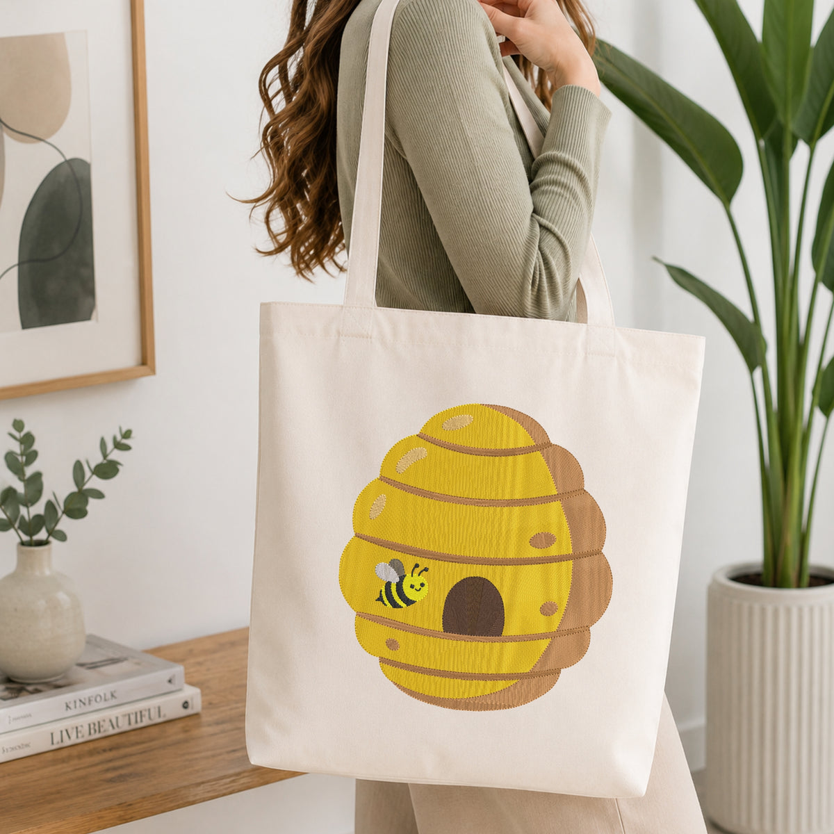 A woman wearing a sage green ribbed sweater carries a cream canvas tote bag with a large yellow beehive and bee graphic on the front, set in a bright, modern room with a wooden table, framed art, and a potted plant.