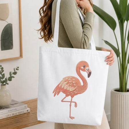 A woman carrying a white canvas tote bag with a large pink flamingo illustration, shown in a bright indoor setting with plants and a wooden table.