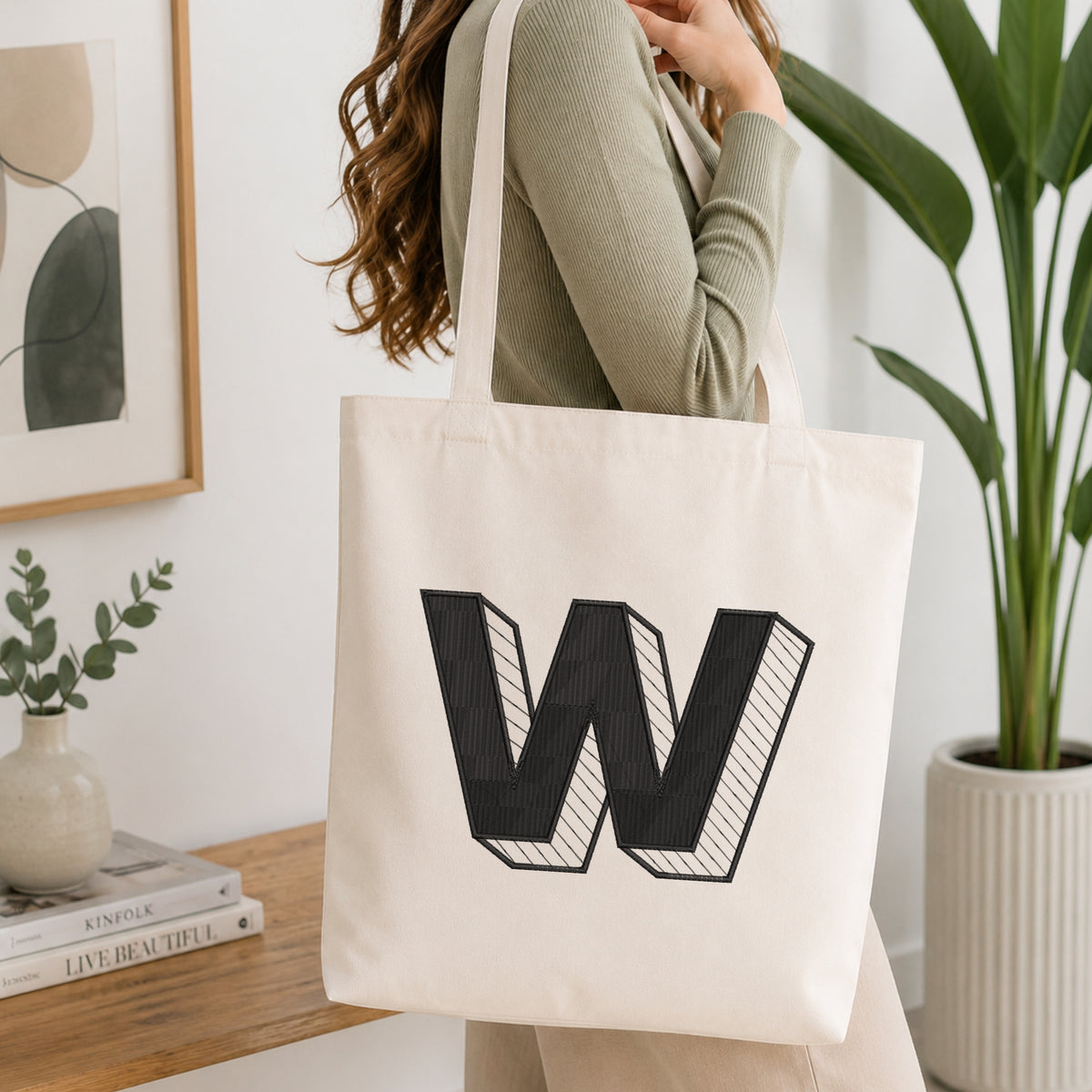 A woman wearing a sage green ribbed long-sleeve top carries a cream canvas tote bag with a large black embroidered letter 'W' on the front, set in a bright, modern room with a wooden console table, framed art, and a potted plant.