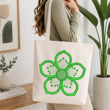 A person wearing a ribbed sage green long-sleeve top carries a cream canvas tote bag with a large green embroidered flower design, set in a bright, modern room with a wooden table, books, and a potted plant.