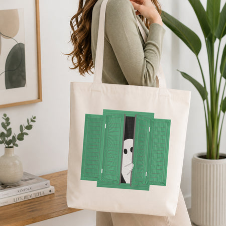 A woman wearing a sage green ribbed sweater carries a cream canvas tote bag with a graphic print of a white ghost peeking out from behind green shutters. The scene is set in a bright, modern room with a wooden table, books, a small plant, and a potted green plant in the background.