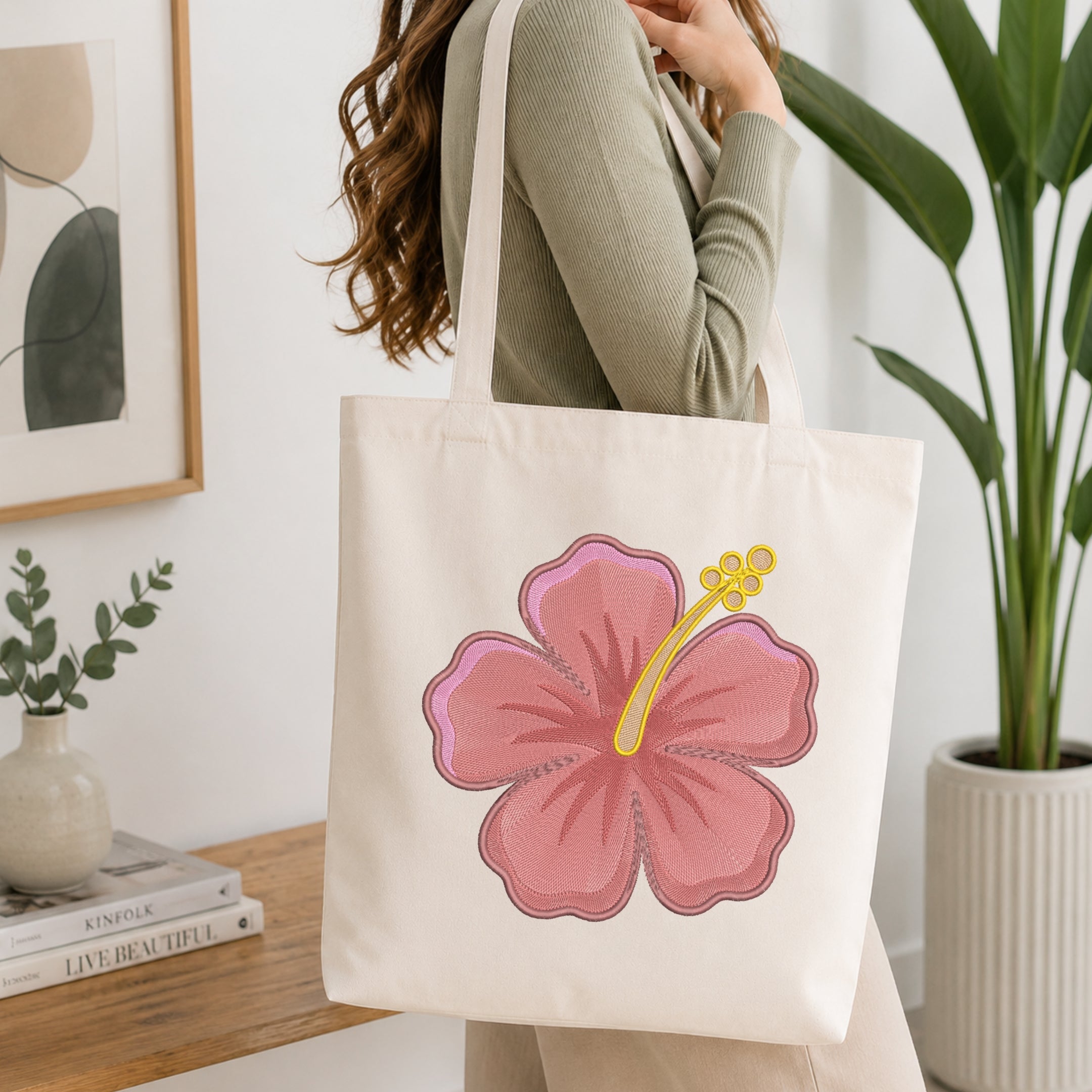 A woman wearing a sage green ribbed long-sleeve top carries a cream canvas tote bag with a large pink hibiscus flower graphic on the front, set in a bright, modern room with a wooden table, books, and a potted plant.
