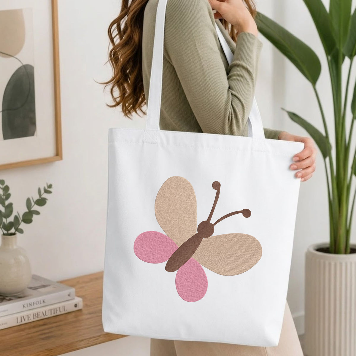 Woman carrying a white natural canvas tote bag featuring a minimalist butterfly machine embroidery design in a bright, modern interior setting