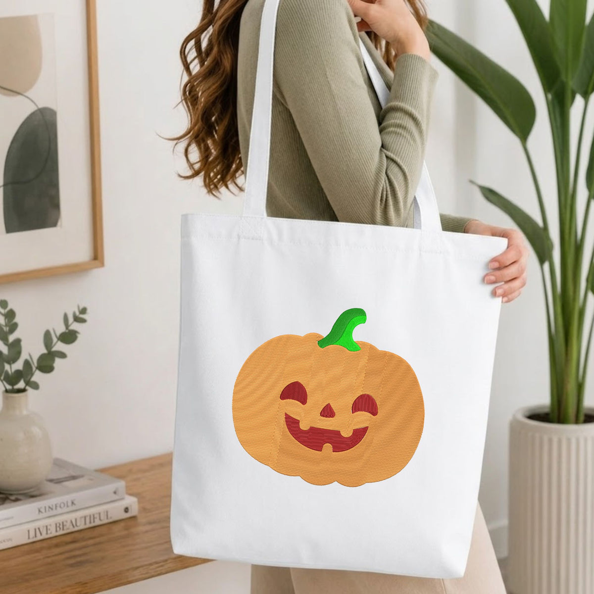 Woman carrying a white natural canvas tote bag featuring an embroidered jack-o'-lantern pumpkin design in a bright, modern interior setting.