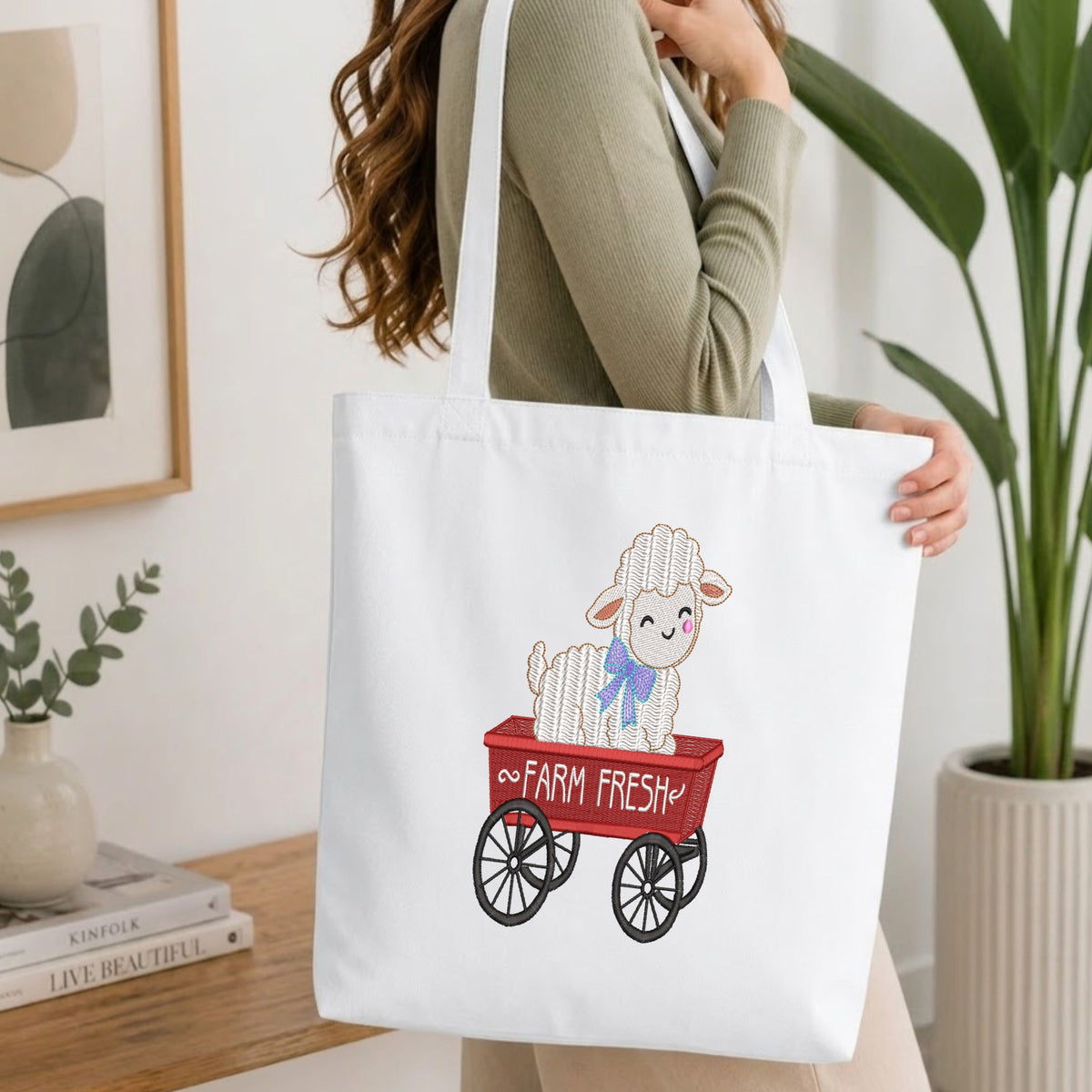  Woman carrying a white natural canvas tote bag featuring an embroidered "Farm Fresh" sheep design in a bright, modern interior setting.   