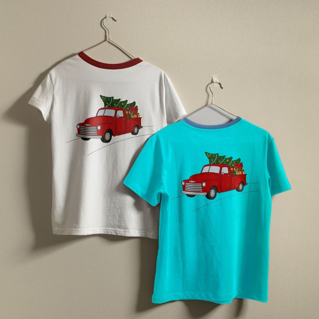 Two short-sleeve crewneck t-shirts hanging on hangers against a beige wall. The left shirt is white with a red collar trim and a graphic of a red pickup truck carrying a Christmas tree.