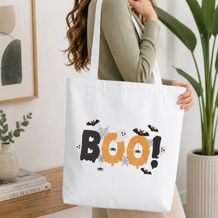 A woman wearing a sage green ribbed long-sleeve top carries a white canvas tote bag with a Halloween-themed 'BOO!' graphic featuring bats, ghosts, spiders, and cobwebs in black, orange, and white.