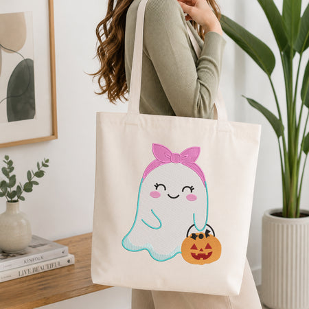 A person wearing a sage green ribbed long-sleeve top carries a cream canvas tote bag with a cute cartoon ghost holding a jack-o'-lantern. The tote bag features a pink bow, a light blue outline, and a smiling face.