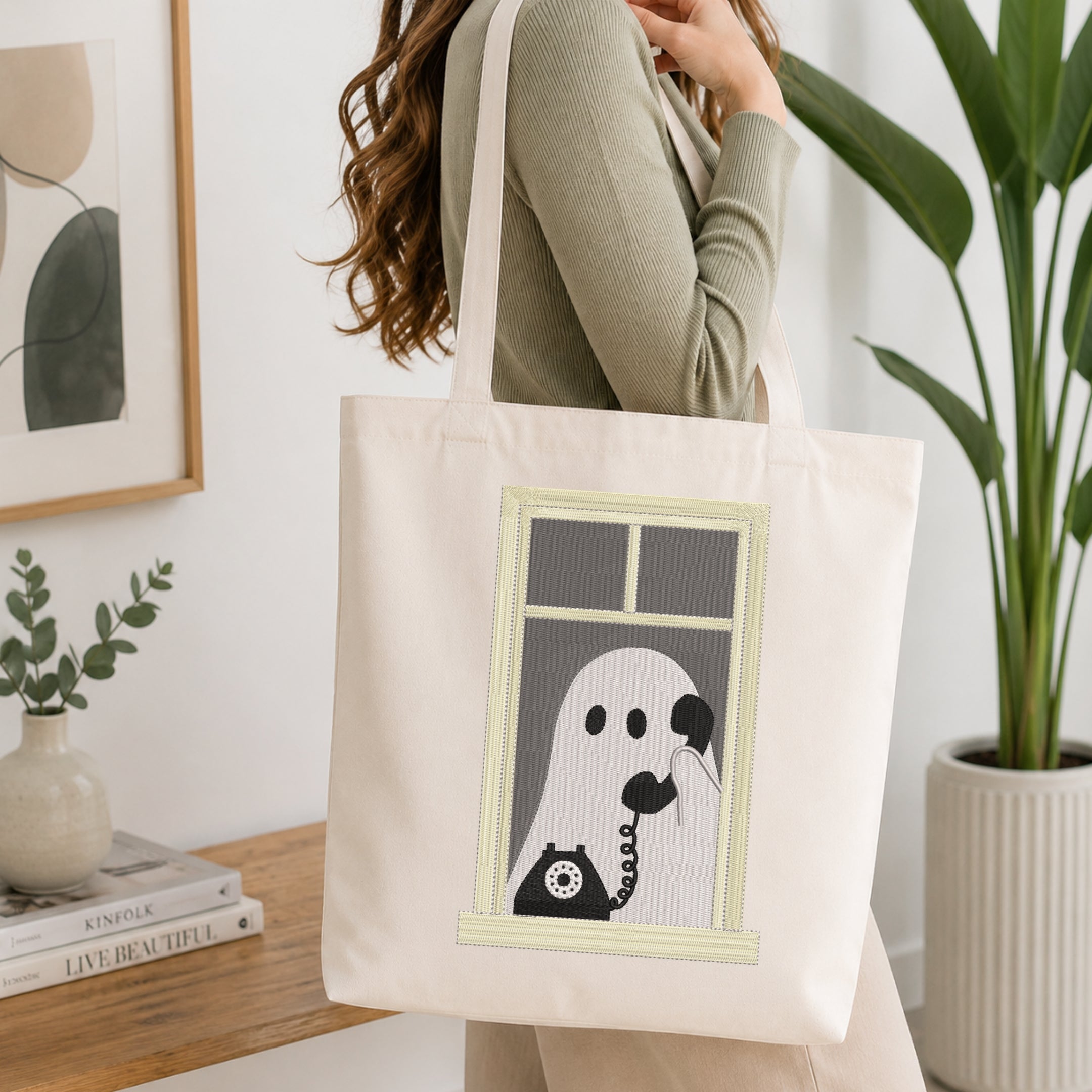 A woman wearing a sage green ribbed sweater carries a cream canvas tote bag with a graphic of a ghost on a telephone looking out a window. The scene is set in a bright, modern room with a wooden table, books, and a potted plant.