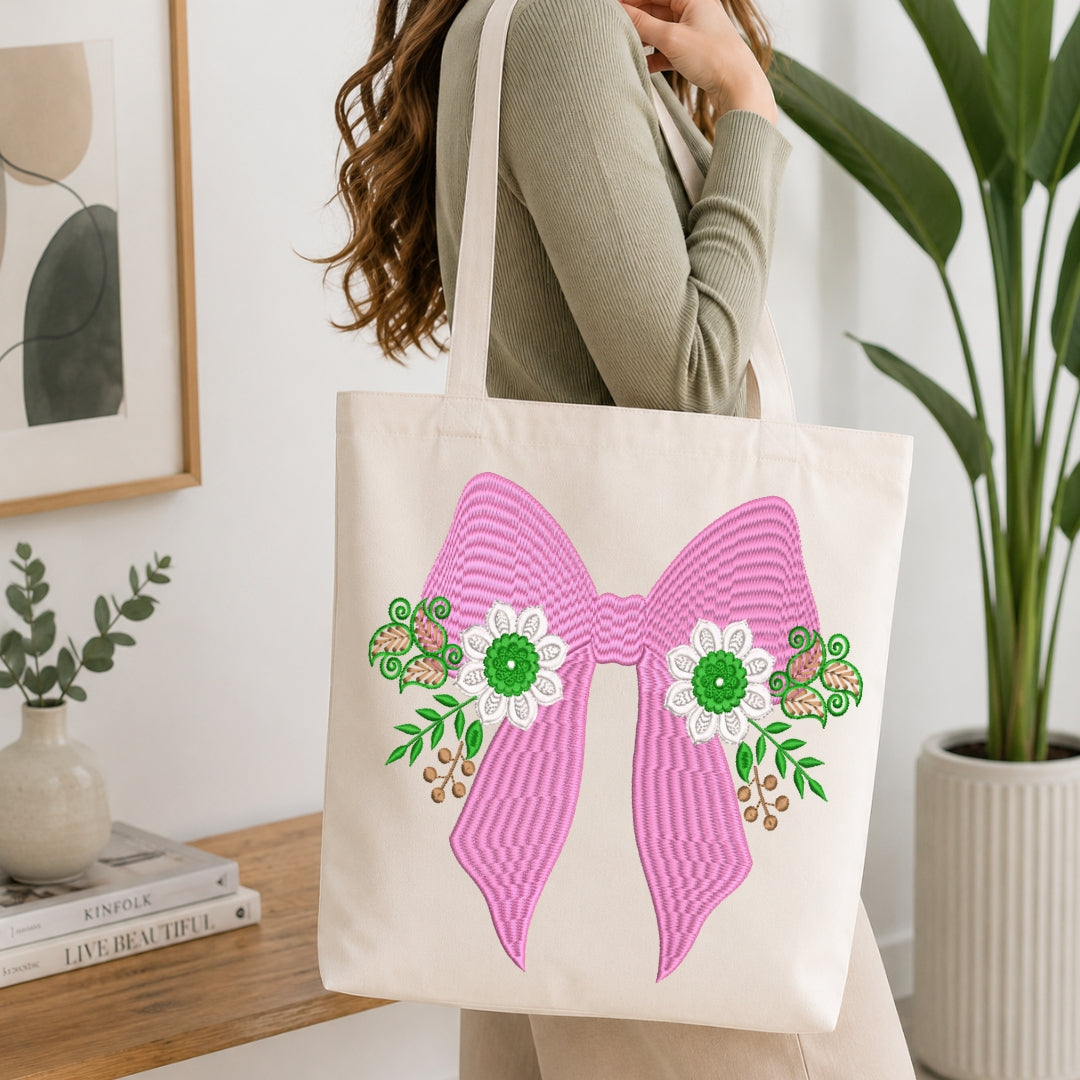 An off-white canvas tote bag with a large pink striped bow and white flowers with green centers, carried by a woman in a ribbed sage green top in a bright, modern room.