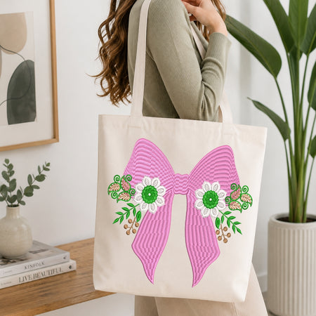 An off-white canvas tote bag with a large pink striped bow and white flowers with green centers, carried by a woman in a ribbed sage green top in a bright, modern room.