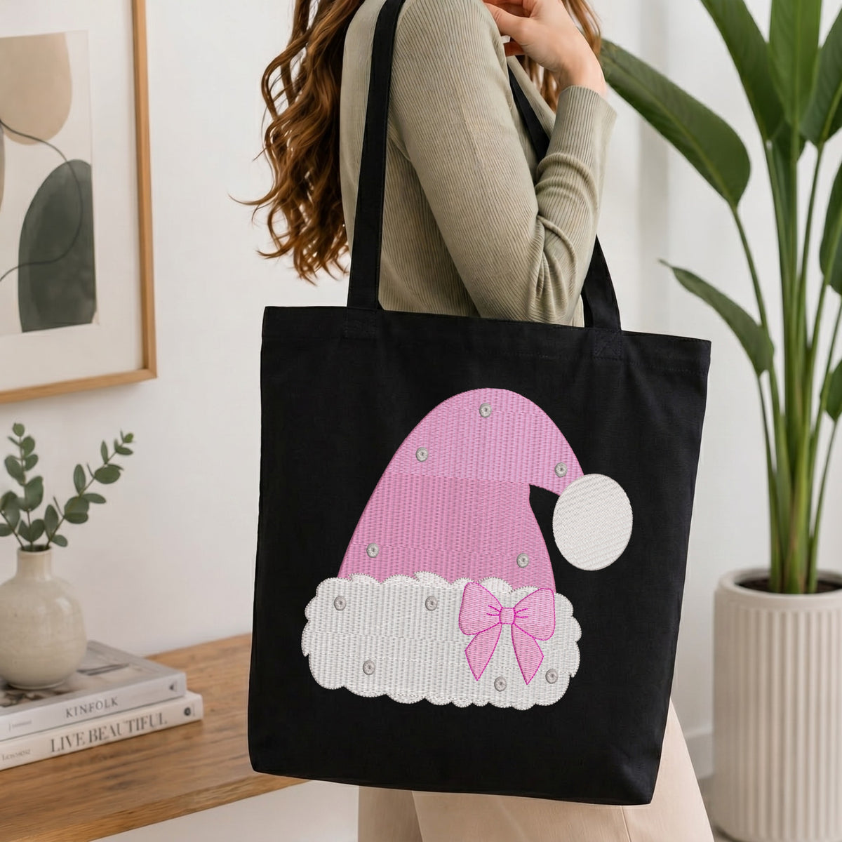 A person wearing a beige ribbed sweater carries a black canvas tote bag featuring a pink and white applique Santa hat design with a pink bow and rhinestone accents, set against a bright, modern indoor background with a wooden table, books, and a potted plant.